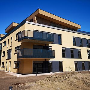 Bei schönstem Wetter präsentiert sich die Wohnhausanlage "Energieffizienter Wohnflair in Wien". Im Vordergrund ist ein Teil des Muliarplatzes zu sehen, der noch in der Gestaltungsphase ist.