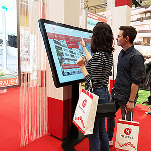 Am Infoscreen informieren sich zwei Messebesucher über die zum Kauf stehenden Eigentumswohnungen in Wien.
