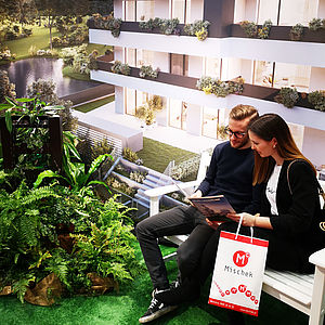Zwei Interessenten auf Wohnungssuche blättern in einem Mischek Folder und sitzen dabei auf einer Parkbank am Messestand.