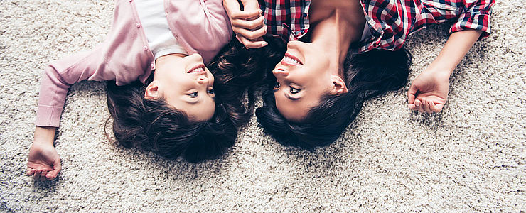 Mutter und Tochter liegen auf einem Teppichboden und lächeln sich an.